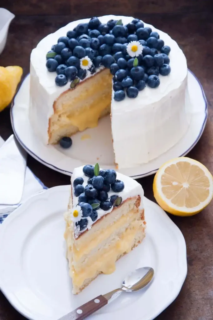 "A beautifully decorated lemon blueberry cake on a cake stand, topped with fresh blueberries and lemon zest