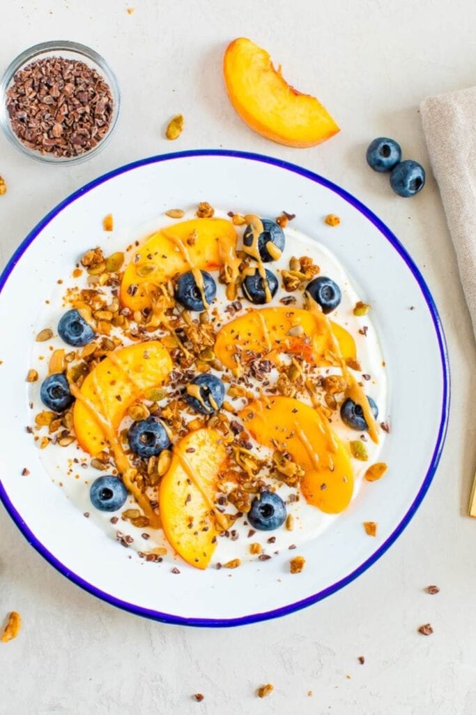 
A bowl of thick Greek yogurt topped with juicy blueberries and sliced peaches, with additional blueberries and peach slices arranged beside the bowl