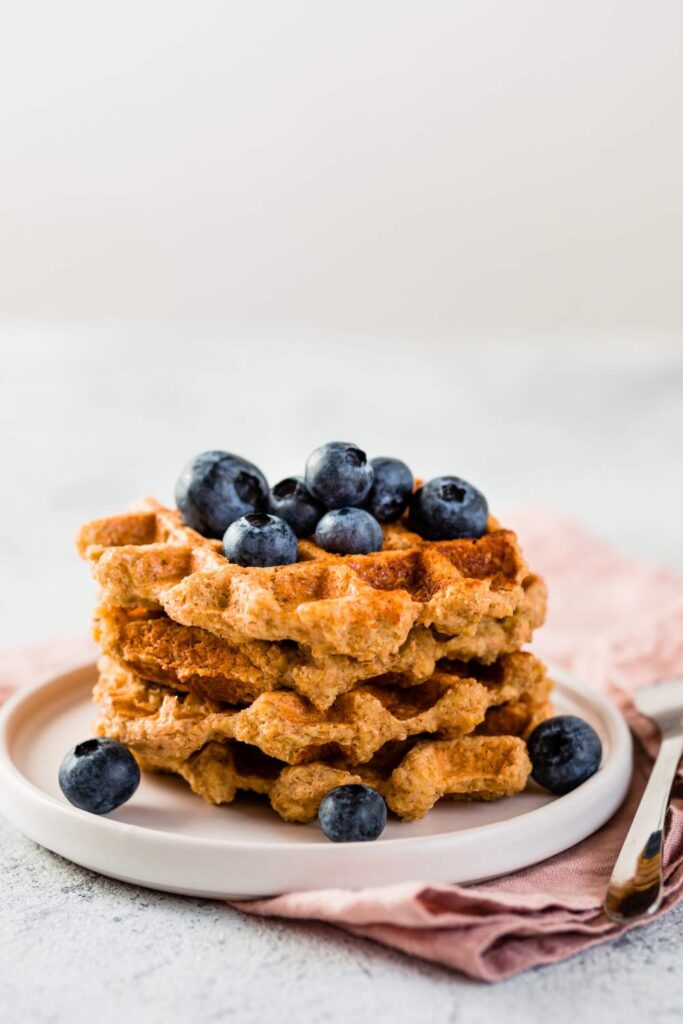 A stack of golden-brown oat waffles served on a white plate, garnished with fresh berries and a drizzle of maple syrup