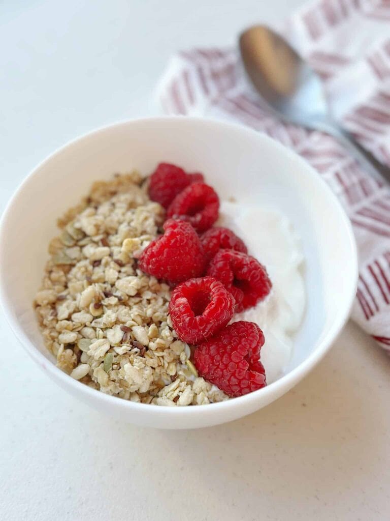 
A Greek yogurt breakfast bowl topped with a dollop of nut butter and an assortment of fresh berries, with a spoon placed beside the bowl on a white marble surface.