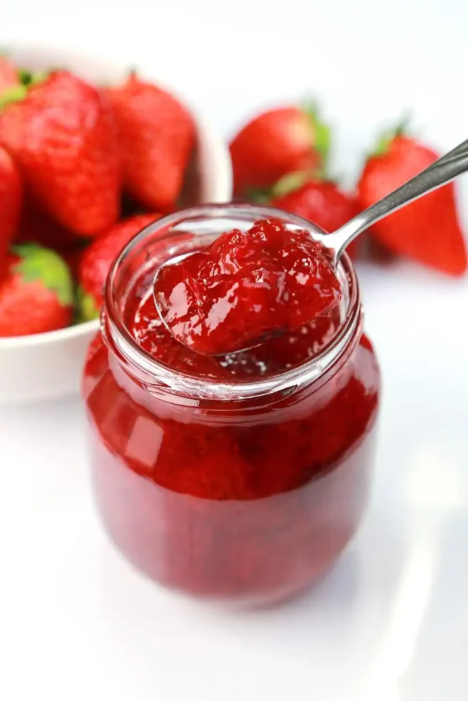 A jar of homemade strawberry jam with fresh strawberries scattered around.