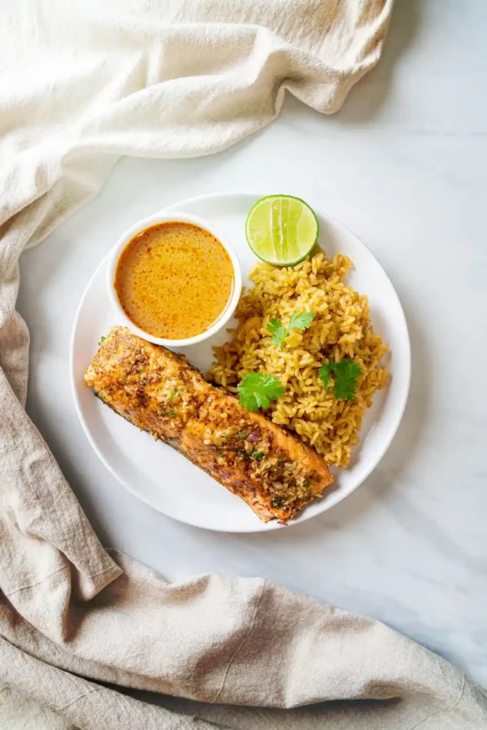 A white plate with a serving of seasoned salmon fillet, yellow spiced rice garnished with cilantro, a halved lime, and a small bowl of orange-hued sauce, set on a light marble surface with a beige cloth napkin.