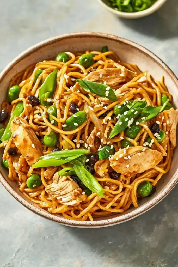 A bowl of stir-fried noodles with chicken, snap peas, and black beans, garnished with sesame seeds and green onions, served on a marble surface with a small bowl of chopped green onions on the side.