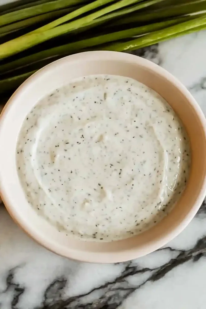 A bowl of creamy dip with visible herbs and spices sprinkled on top, placed on a marble surface. Green onions or chives are arranged in the background.