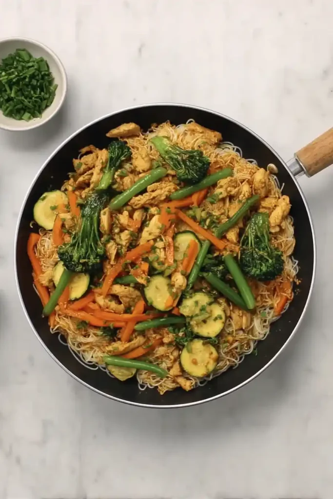 A pan of stir-fried noodles with chicken, broccoli, carrots, zucchini, and green beans, garnished with chopped green onions, on a marble surface, with a small bowl of additional green onions on the side.