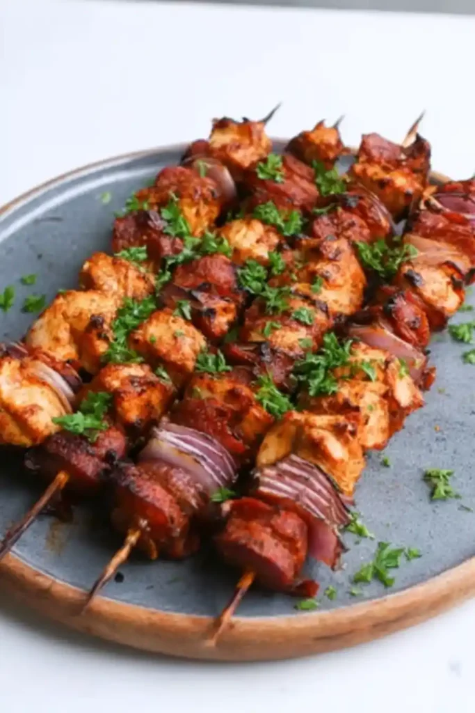 Six skewers of grilled chicken and sausage kebabs, interspersed with red onion slices, arranged on a round gray plate with a wooden rim.