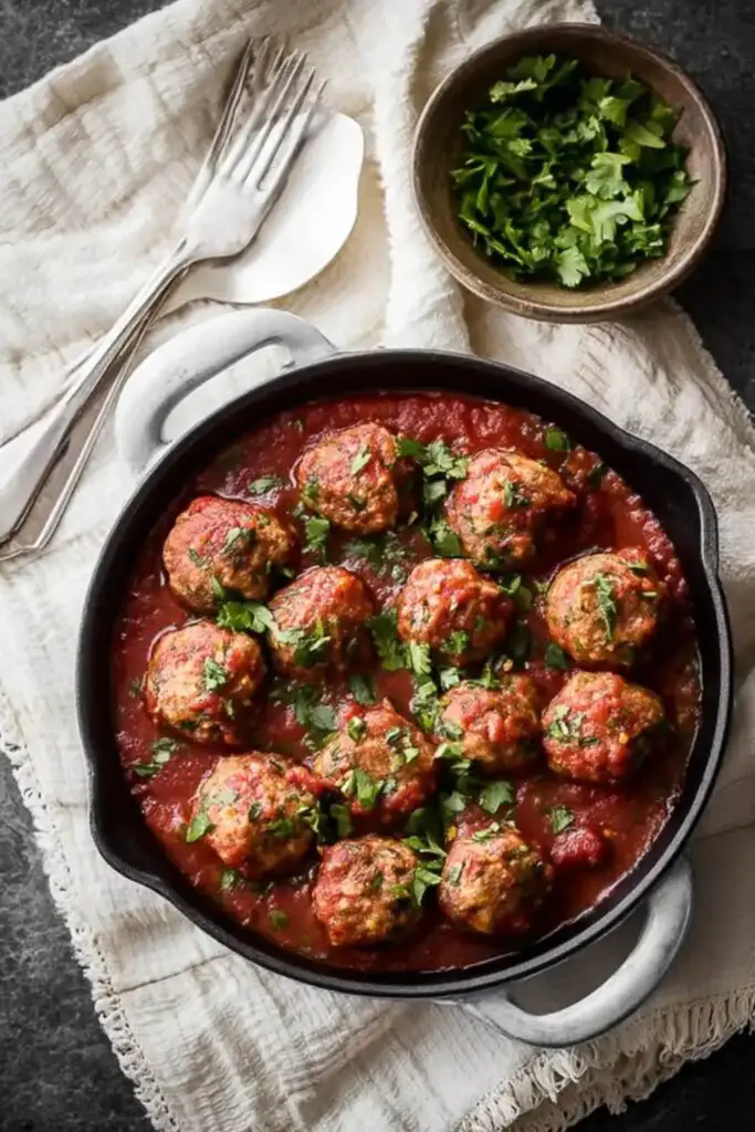 A rustic black cast-iron pan filled with meatballs simmered in a rich tomato sauce, garnished with fresh parsley. The pan is placed on a textured white napkin .