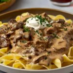 A plate of egg noodles topped with creamy beef stroganoff, garnished with a dollop of sour cream and chopped parsley.