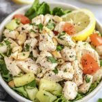 A white bowl of salad with leafy greens, shredded chicken, diced tomatoes, cucumbers, lemon slices, and chopped parsley, set on a marble surface with a gray cloth napkin and lemon wedges in the background.