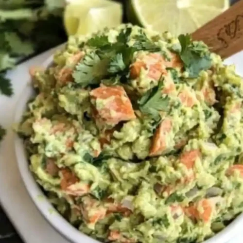 A white bowl of chunky guacamole with diced tomatoes, onions, and chopped cilantro, garnished with lime slices and a sprig of cilantro, served with a wooden spoon on a dark marble surface.