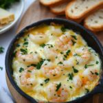 Creamy baked shrimp topped with melted cheese and fresh parsley, served in a skillet with slices of crusty bread on the side.