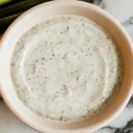 A bowl of creamy dip with visible herbs and spices sprinkled on top, placed on a marble surface. Green onions or chives are arranged in the background.