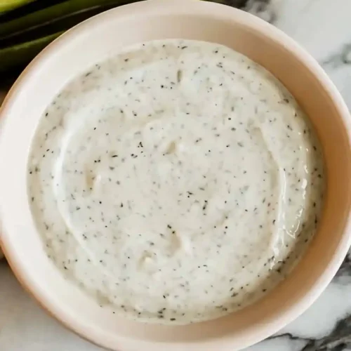 A bowl of creamy dip with visible herbs and spices sprinkled on top, placed on a marble surface. Green onions or chives are arranged in the background.