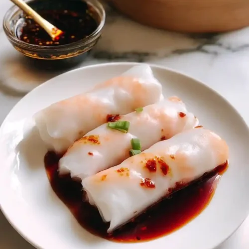 Three shrimp rice noodle rolls (Cheung Fun) served on a white plate, topped with soy-based sauce and garnished with chopped green onions and chili oil, with a bowl of dipping sauce and a bamboo steamer in the background.