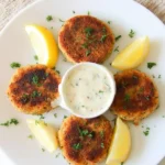 A white plate featuring four golden salmon patties arranged in a circular pattern, garnished with fresh parsley. In the center, a small bowl of creamy dipping sauce is placed, with lemon wedges and additional parsley scattered around the plate.